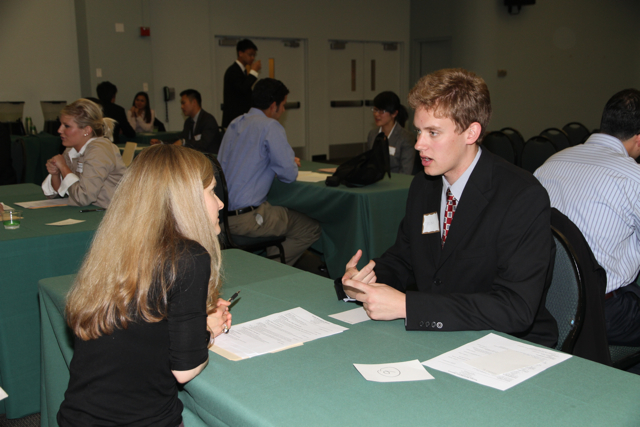 Two people speaking across a table with application papers, with other interview conversations behind them.