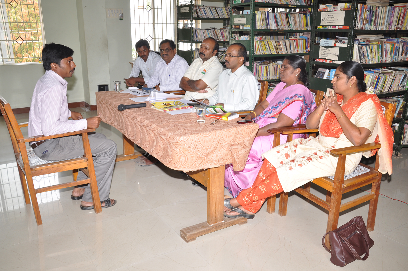 A seated candidate faces a panel across a table in a room lined with bookshelves.