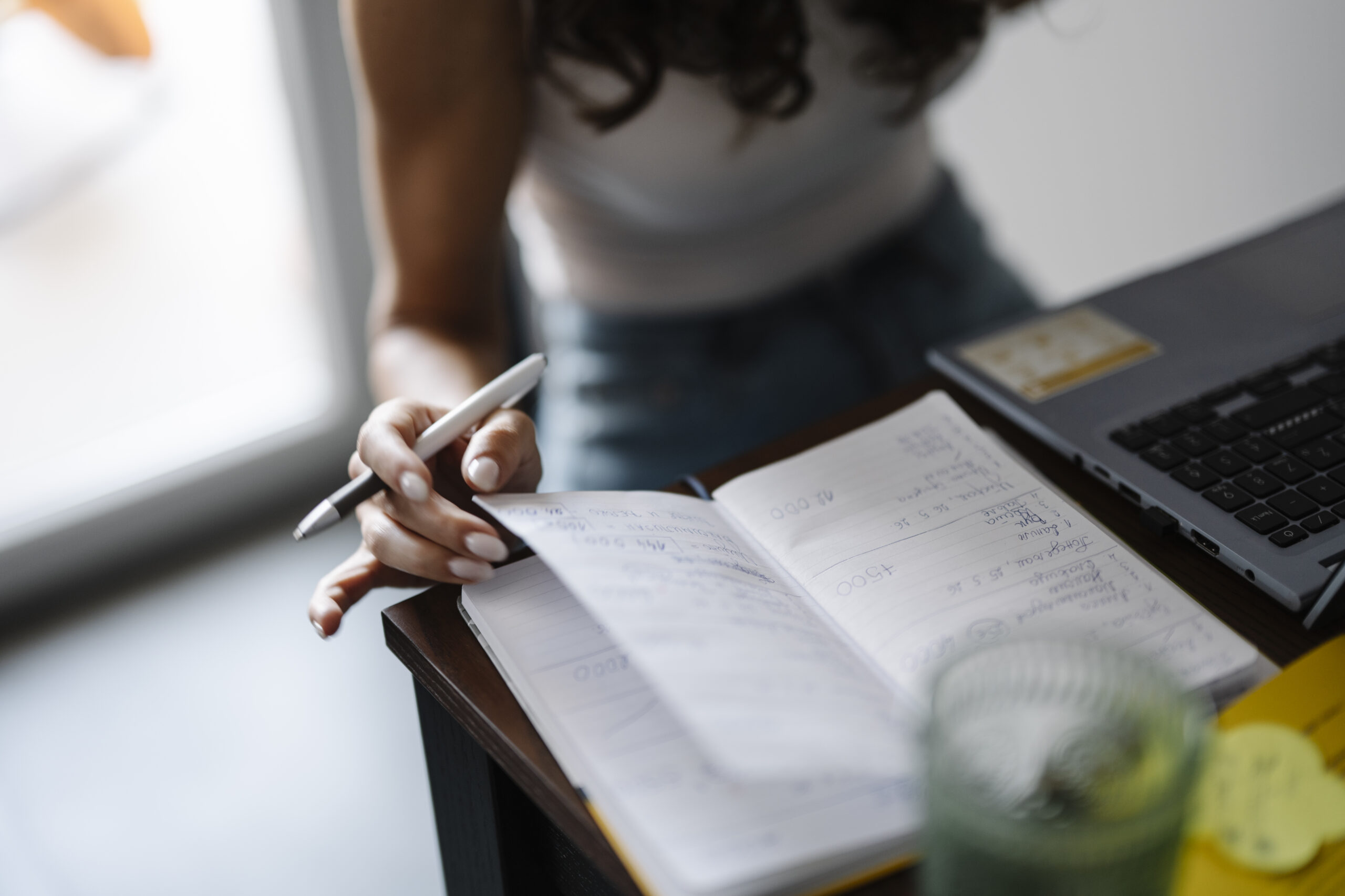 Person holding a pen over a notebook beside an open laptop and a glass of water.